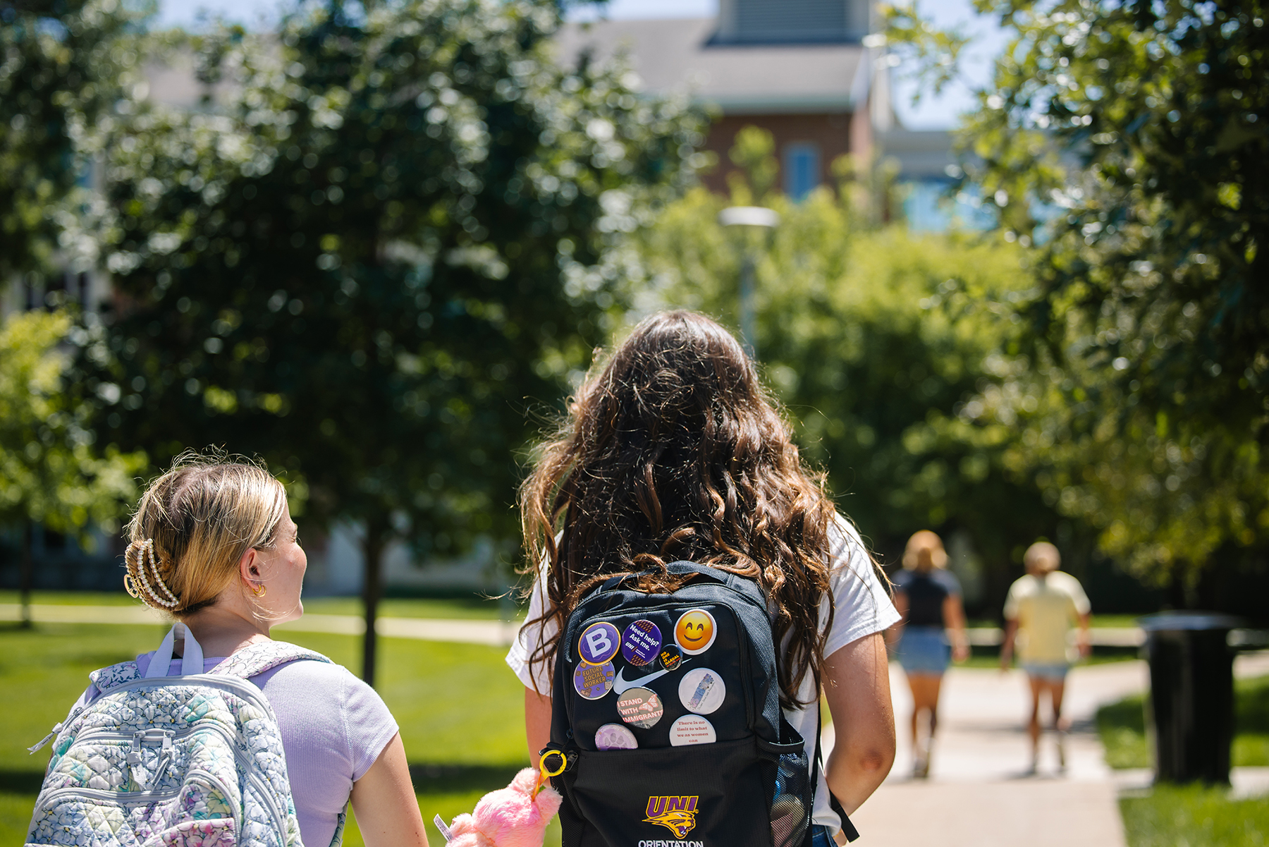 Students wear backpacks covered in statement pins and a UNI logo patch while walking on campus
