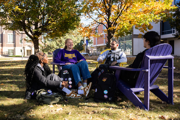 Group of students sitting outside on the ground and in Adirondack chairs
