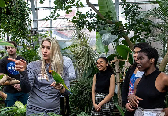 A group of students smiling as an instructor in a greenhouse talks while holding a tropical bird