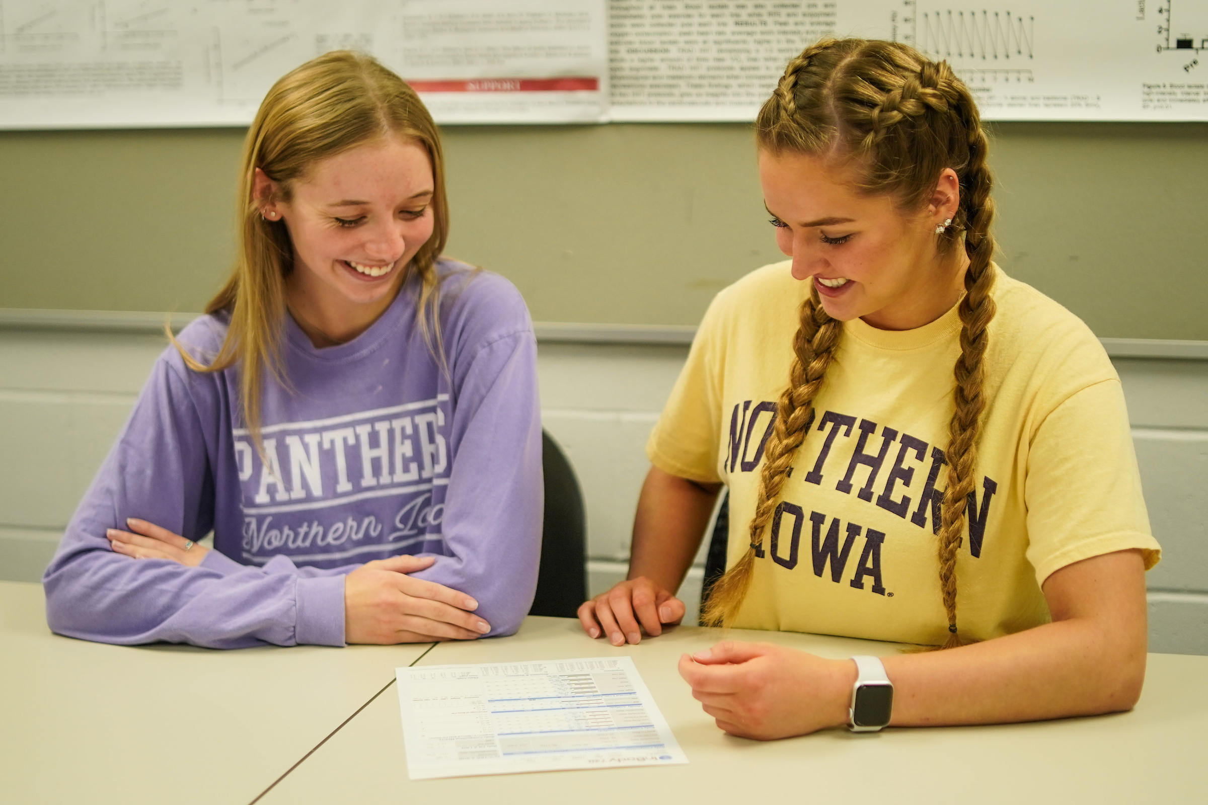 Two students sitting at desk, smiling and looking at a paper