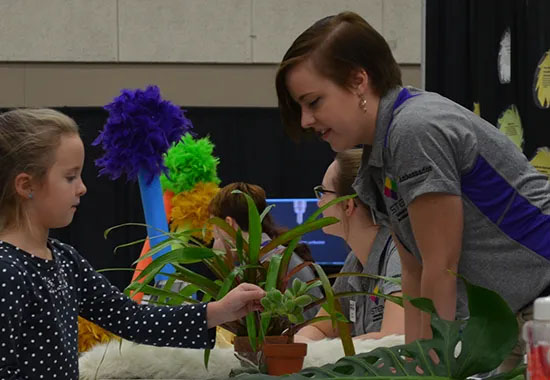An elementary student interacts with a plant as a college student talks to her