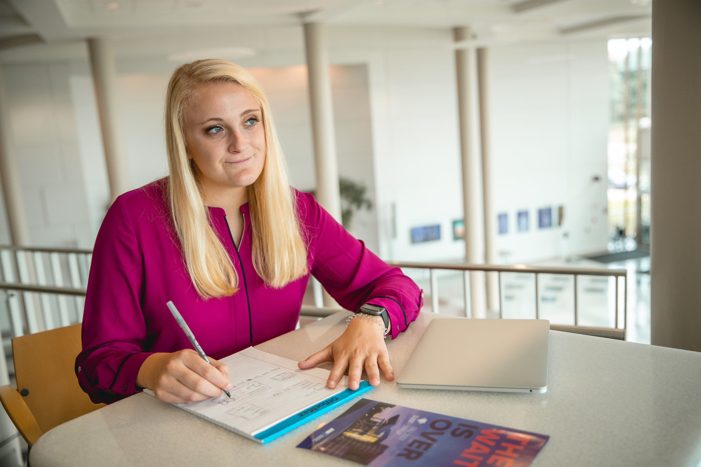 Student sitting at table while looking off into the distance, pen ready to write in notebook